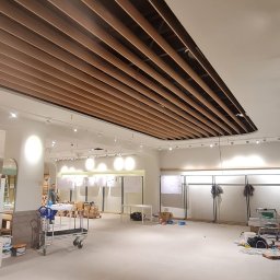 WEBEKO Maciej Paśnik - Wnętrze sklepu w trakcie prac wykończeniowych, widoczny montaż drewnianej podwieszanej konstrukcji sufitowej z oświetleniem punktowym, przygotowane ściany i podłogi, wózek transportowy...