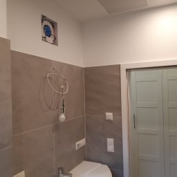 WEBEKO Maciej Paśnik - Niedokończona łazienka z szarą glazurą, białą umywalką i wiszącą żarówką. Widoczny otwór w ścianie z wentylatorem.