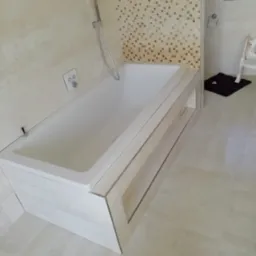 Nowoczesna biała wanna obudowana płytkami imitującymi kamień, z mozaiką w tle, chromowana słuchawka prysznicowa i armatura, jasne płytki podłogowe, fragment czarnej wycieraczki i krzesełka w rogu.