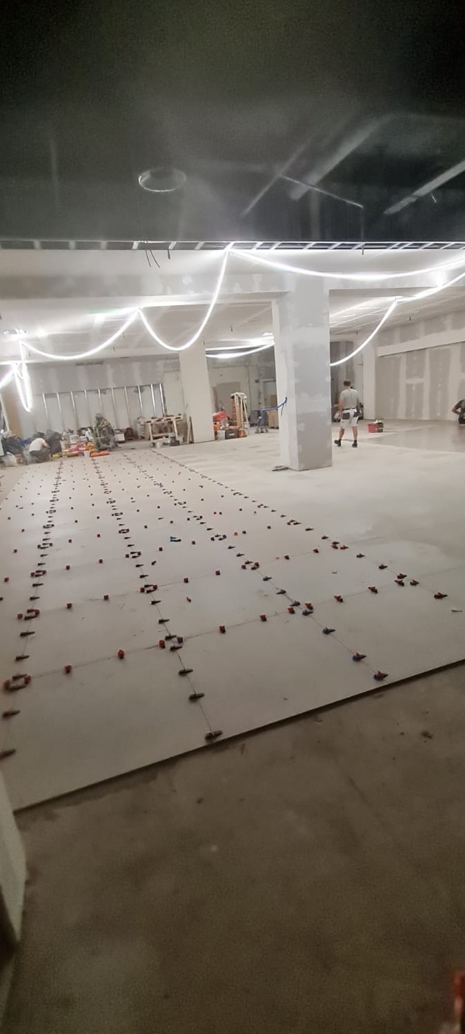 Przygotowanie do układania płytek w dużym pomieszczeniu, widoczne poziomowanie płytek za pomocą klipsów i klinów, w tle oświetlenie budowlane i pracownicy.