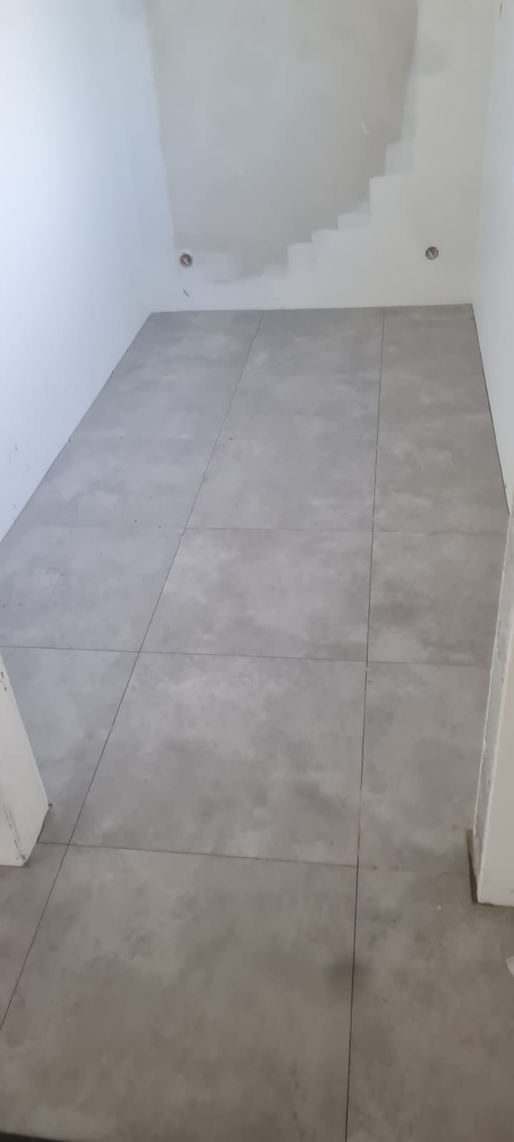 Szare, matowe płytki podłogowe ułożone w pomieszczeniu w trakcie prac wykończeniowych, widoczne niedoskonałości na ścianach i brak listew przypodłogowych.