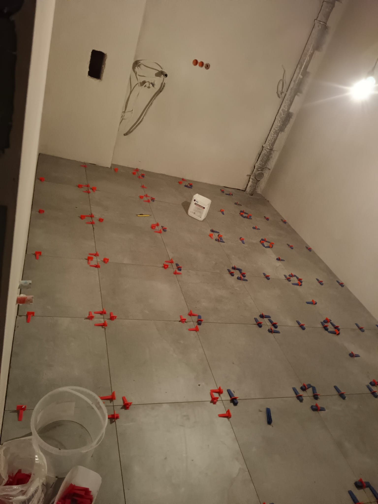 Szare płytki podłogowe w trakcie układania, z czerwonymi i niebieskimi krzyżykami dystansowymi, widoczny biały pojemnik z zaprawą, instalacja elektryczna na suficie w tle.