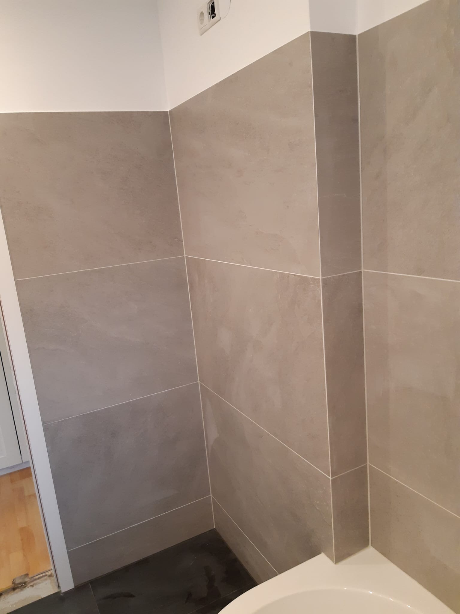 Ściana łazienki wyłożona szarymi płytkami ceramicznymi z jasną fugą, widoczny fragment podłogi z ciemnych płytek oraz narożnik wanny.