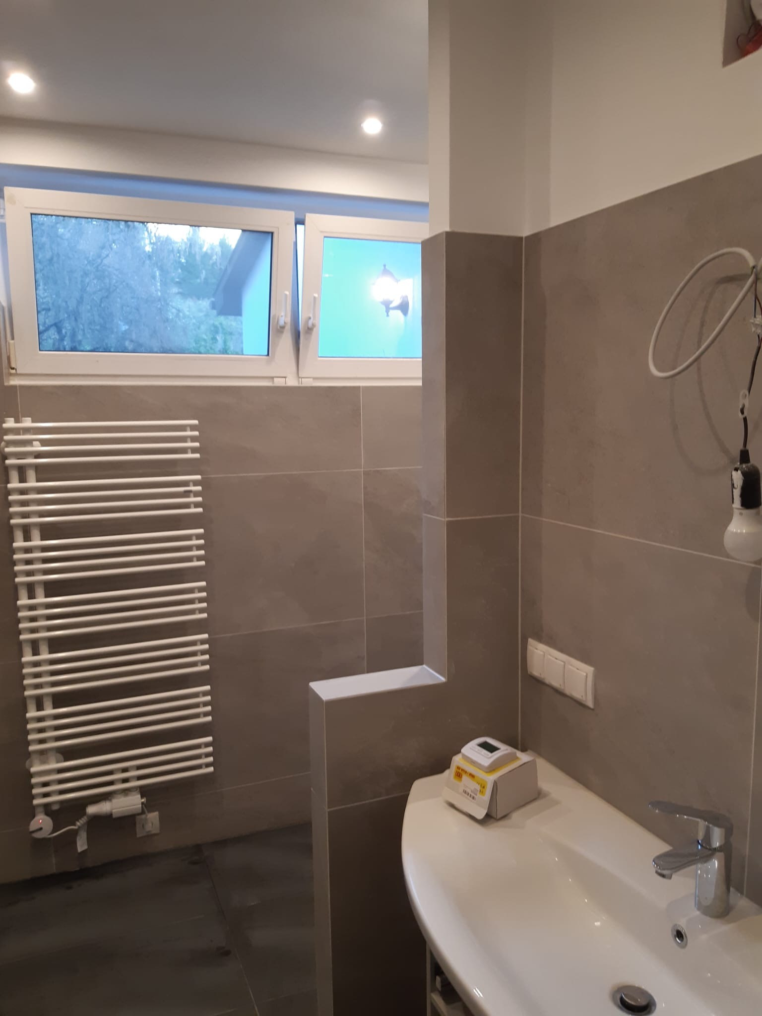 Nowoczesna łazienka z szarymi płytkami, białym grzejnikiem drabinkowym, umywalką i oknem z widokiem na zewnątrz. Na umywalce pudełko z urządzeniem pomiarowym.