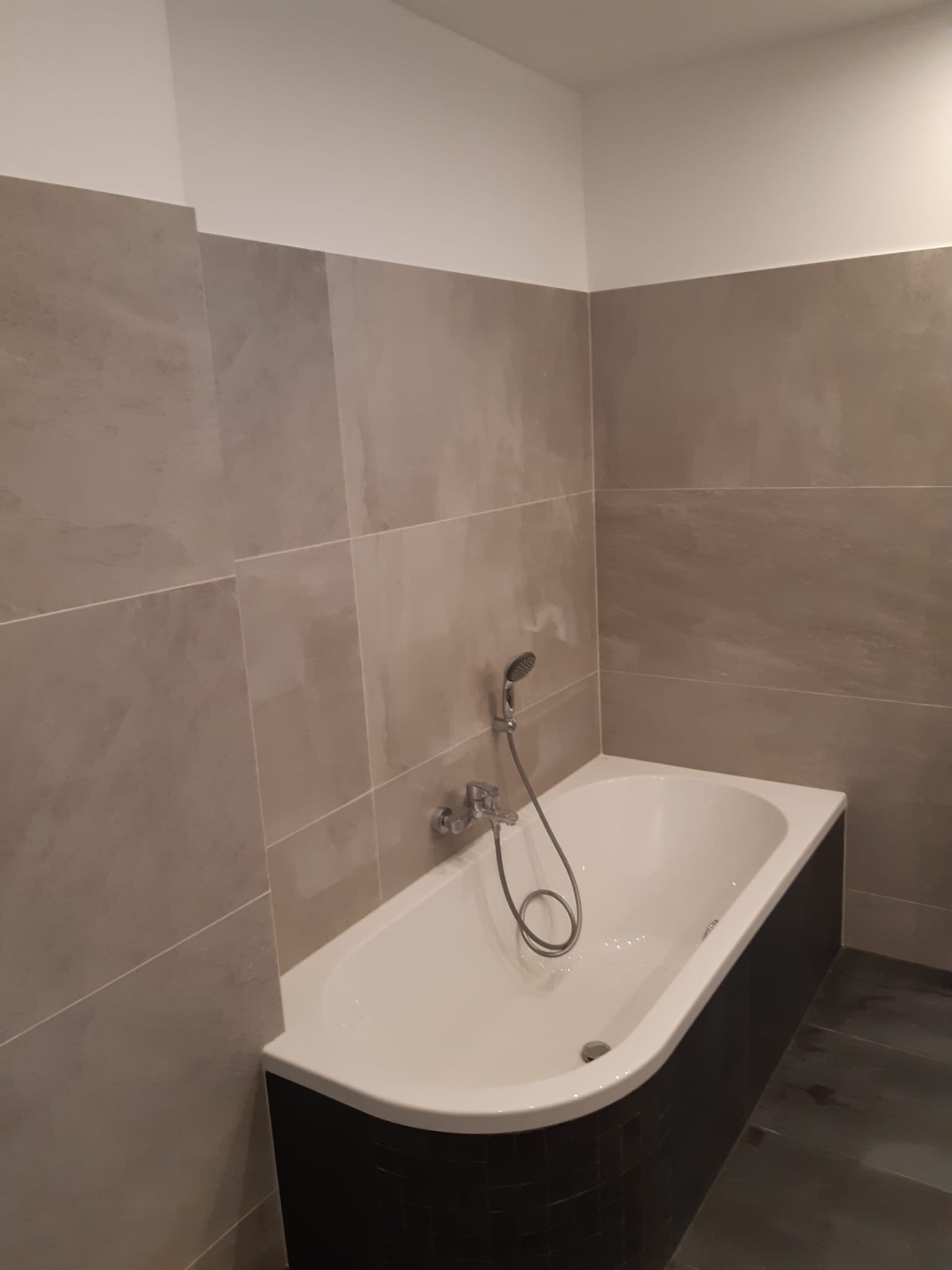 Nowoczesna łazienka z beżowymi płytkami imitującymi kamień na ścianach, białą wanną z czarną obudową i chromowaną słuchawką prysznicową.