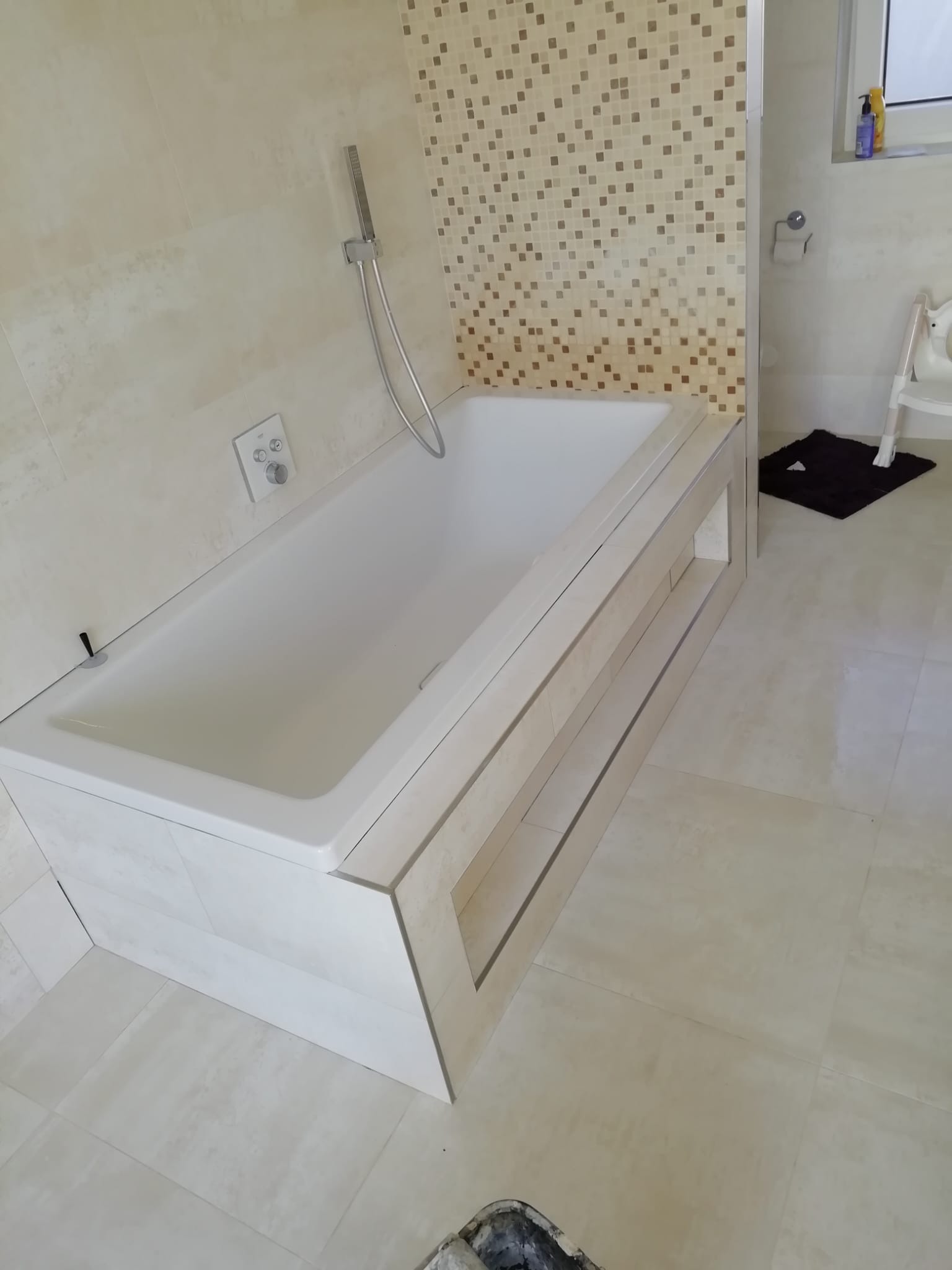 Nowoczesna biała wanna obudowana płytkami imitującymi kamień, z mozaiką w tle, chromowana słuchawka prysznicowa i armatura, jasne płytki podłogowe, fragment czarnej wycieraczki i krzesełka w rogu.