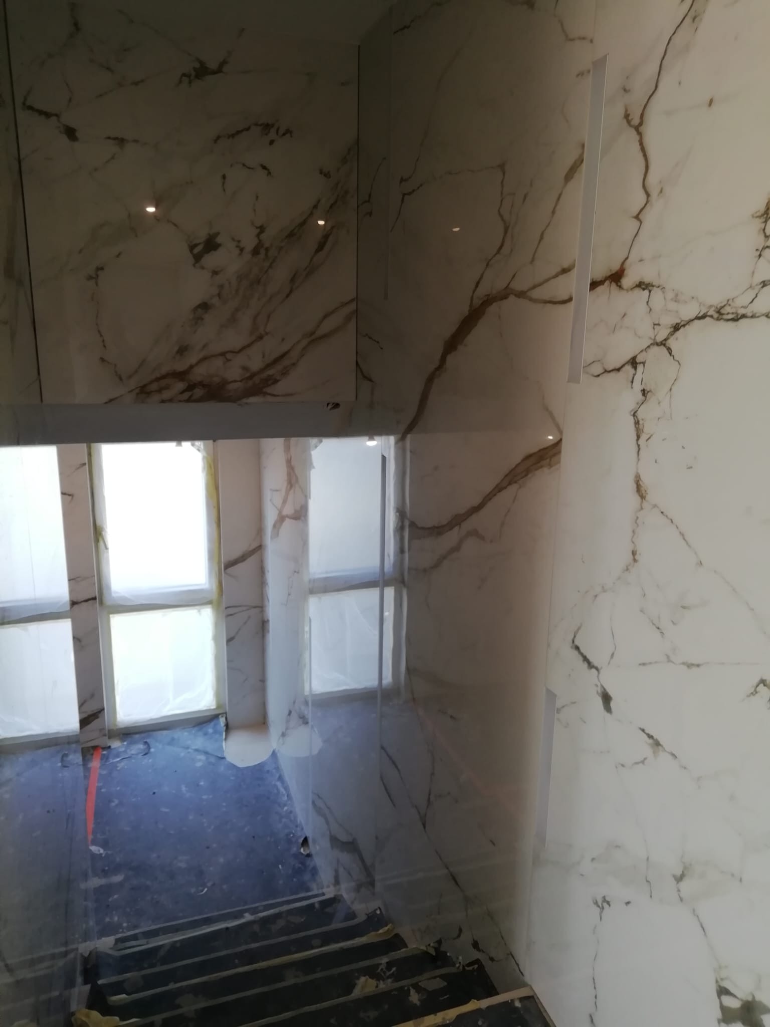Klatka schodowa wykończona białym marmurem z brązowymi żyłami, okna zabezpieczone folią malarską, widoczne schody tymczasowe.