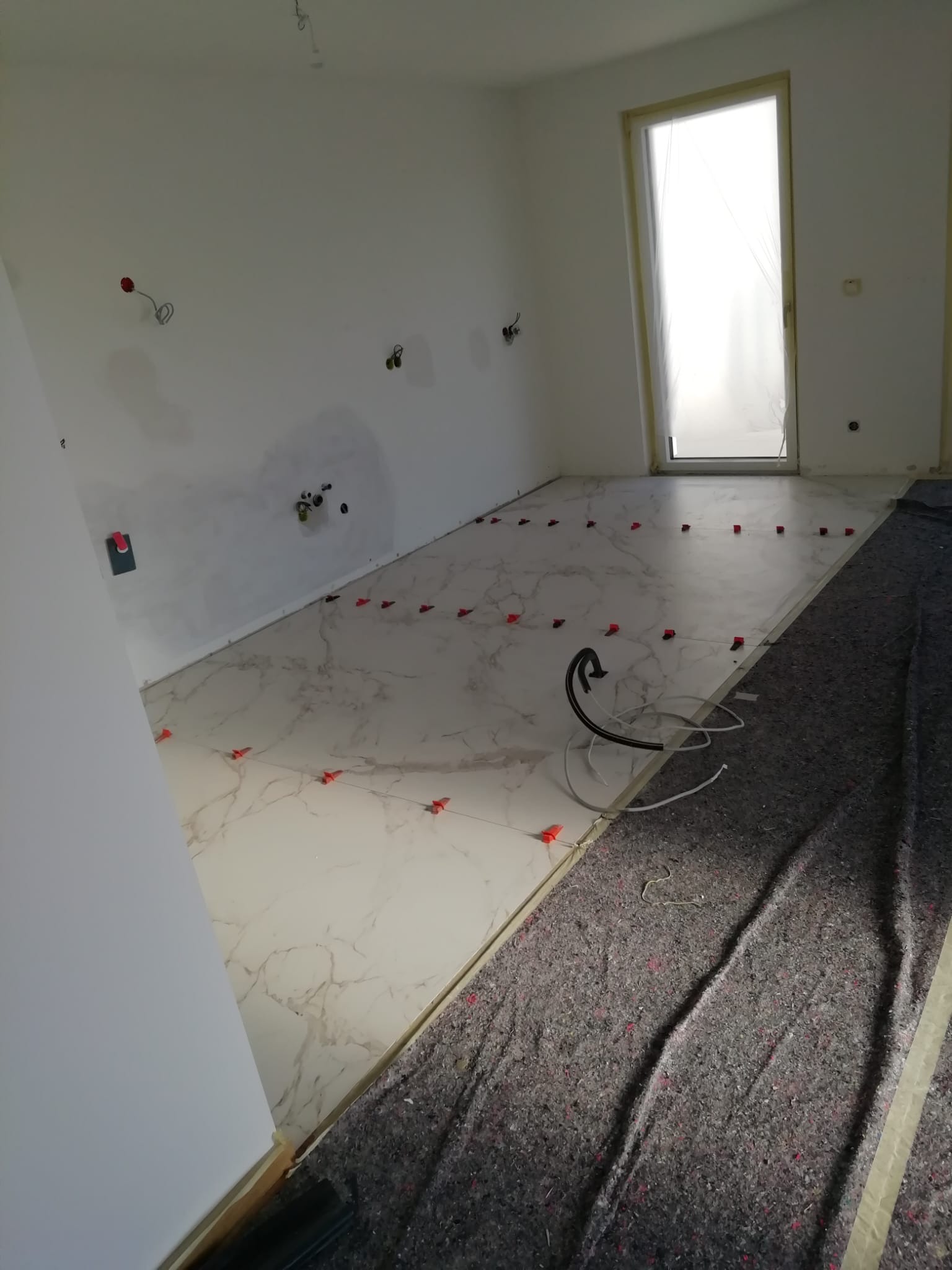 Podłoga z płytek imitujących marmur w trakcie układania, z widocznymi klipsami poziomującymi, zabezpieczona flizeliną malarską przy ścianach, okno zabezpieczone folią.