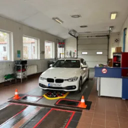 Białe BMW na stanowisku diagnostycznym w warsztacie; widoczne kanały najazdowe z kratami, pomarańczowe pachołki i tablica z wynikami pomiarów.
