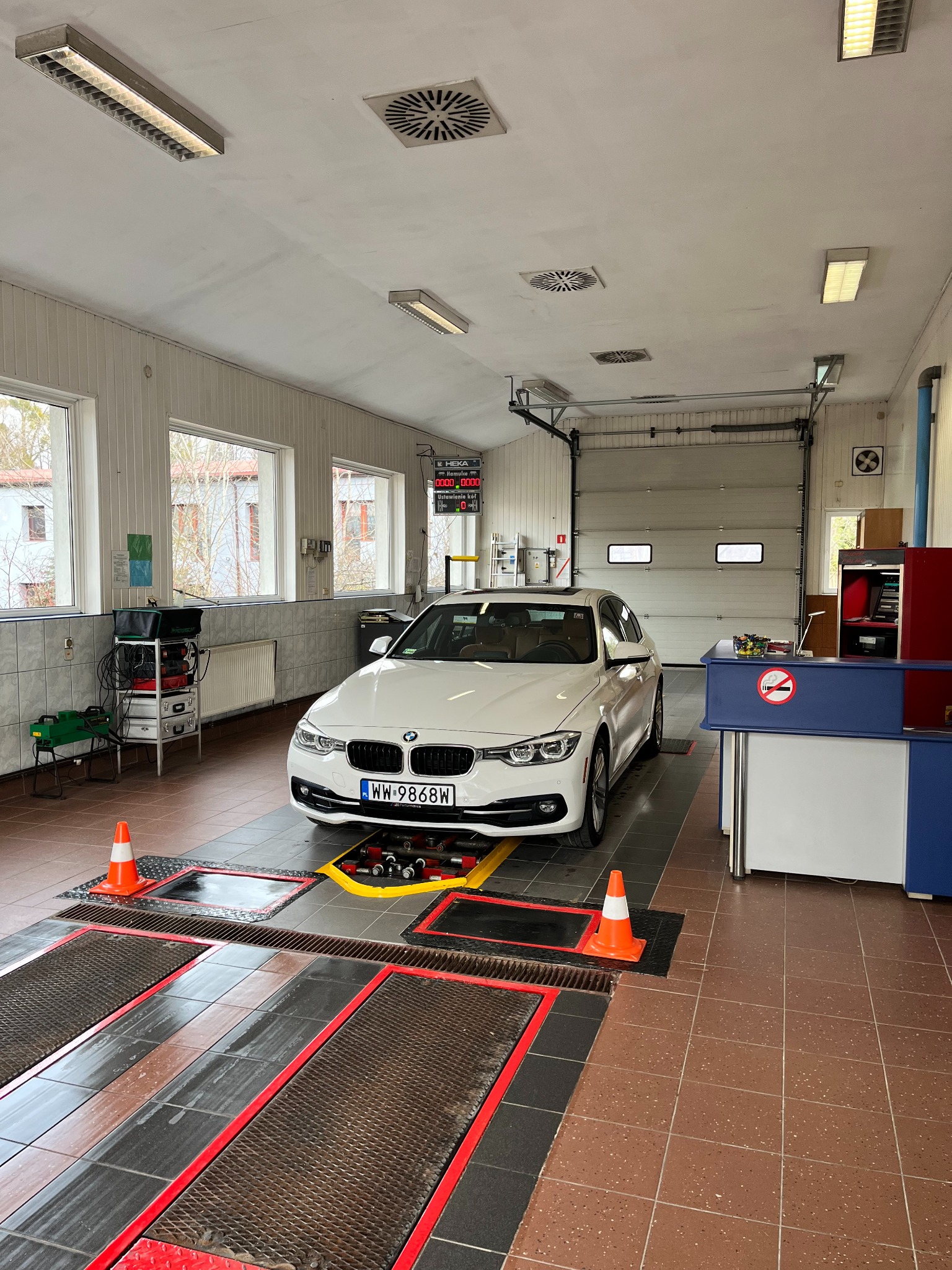 Białe BMW na stanowisku diagnostycznym w warsztacie; widoczne kanały najazdowe z kratami, pomarańczowe pachołki i tablica z wynikami pomiarów.