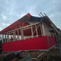Konstrukcja drewniana domu w trakcie budowy, widoczna więźba dachowa z częściowo rozłożoną membraną, czerwone panele na ścianach szczytowych, pochmurne niebo w tle, widoczne narzędzia i materiały...