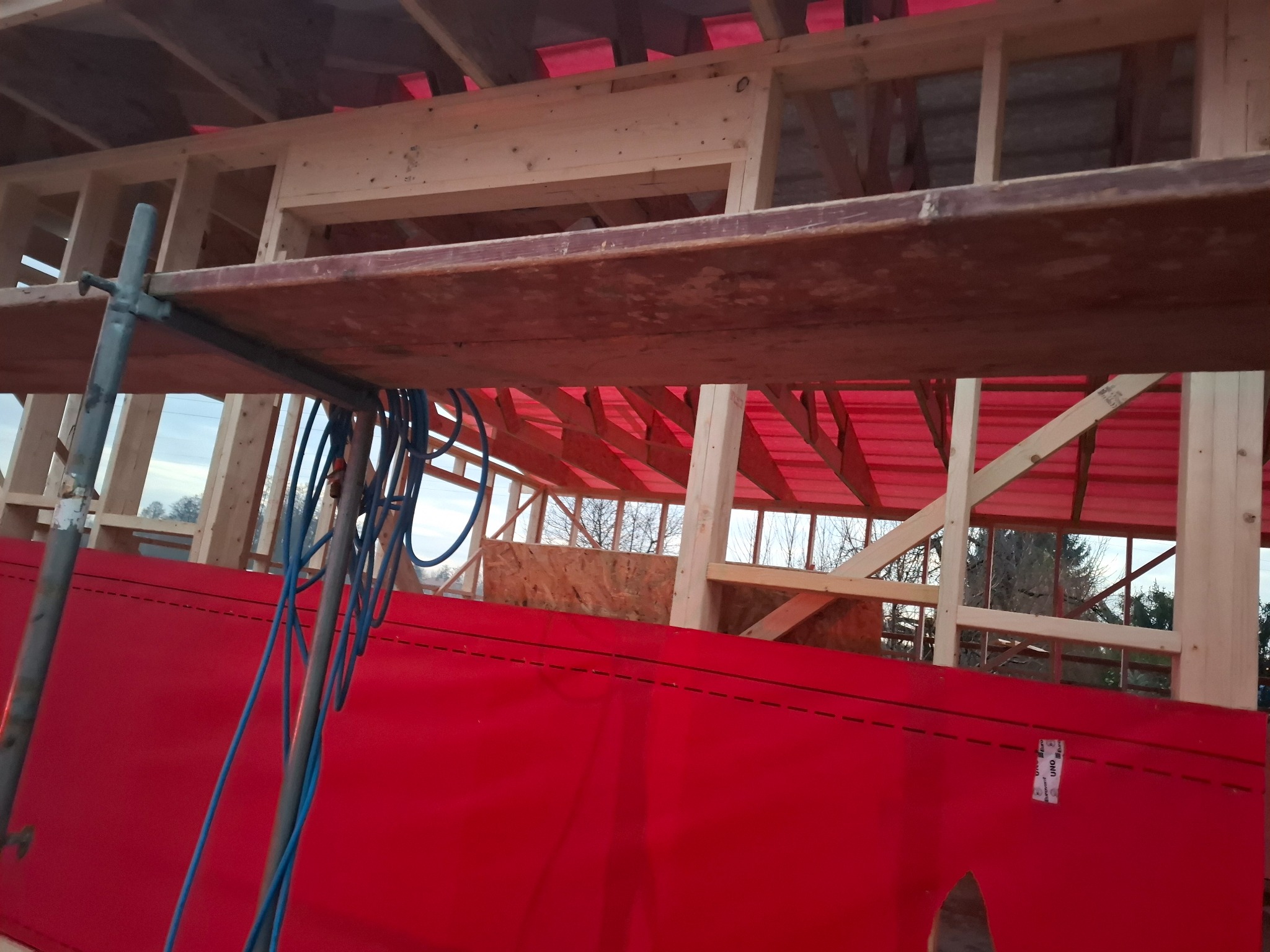 Wnętrze konstrukcji dachu w trakcie budowy, widoczna więźba dachowa, czerwona membrana dachowa oraz fragment rusztowania z niebieskimi kablami.