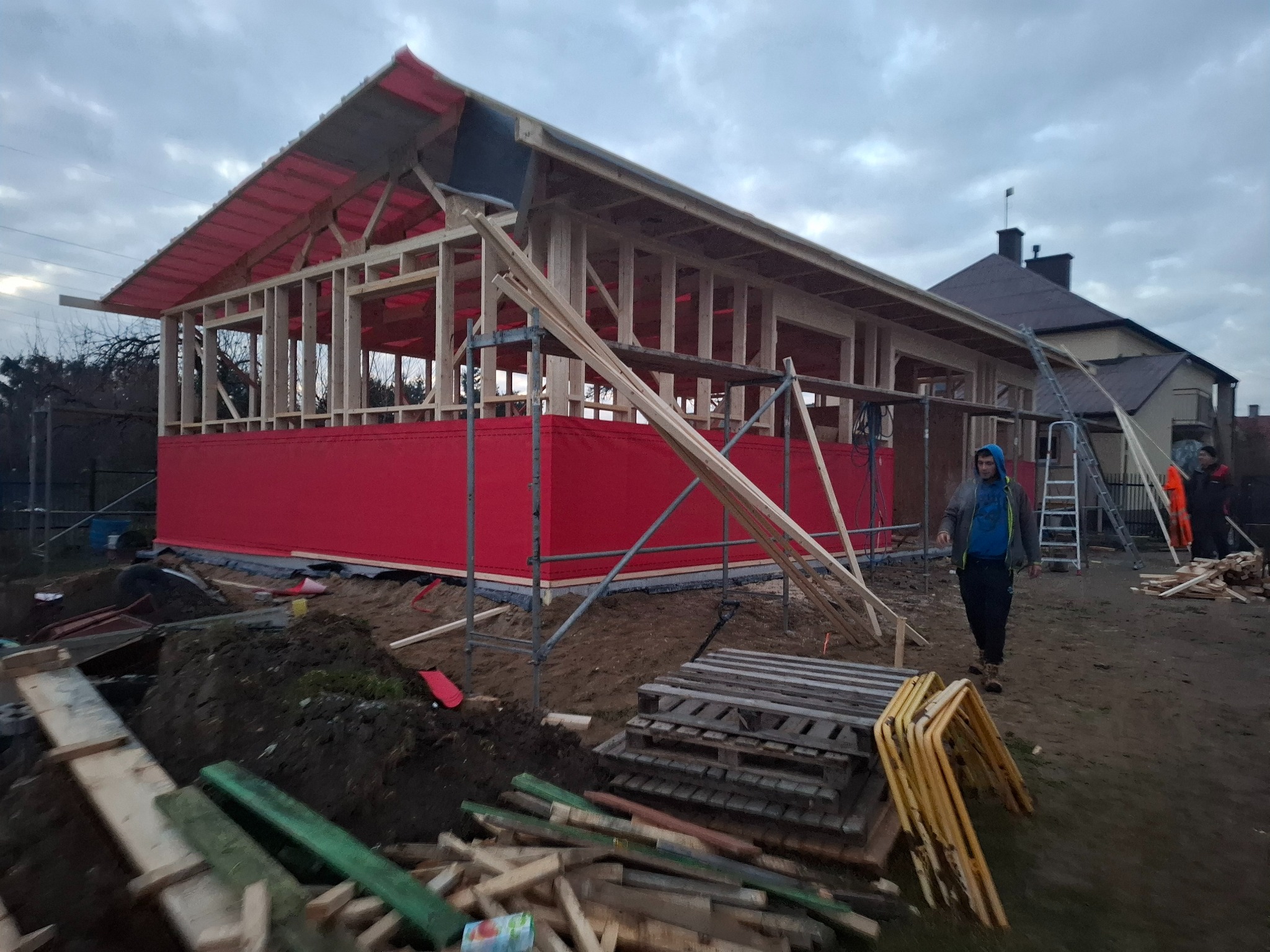 Konstrukcja drewniana budynku z częściowo zamontowaną czerwoną membraną dachową i ścianami, widoczna więźba dachowa, w tle dom mieszkalny, na pierwszym planie stosy drewna i palety.