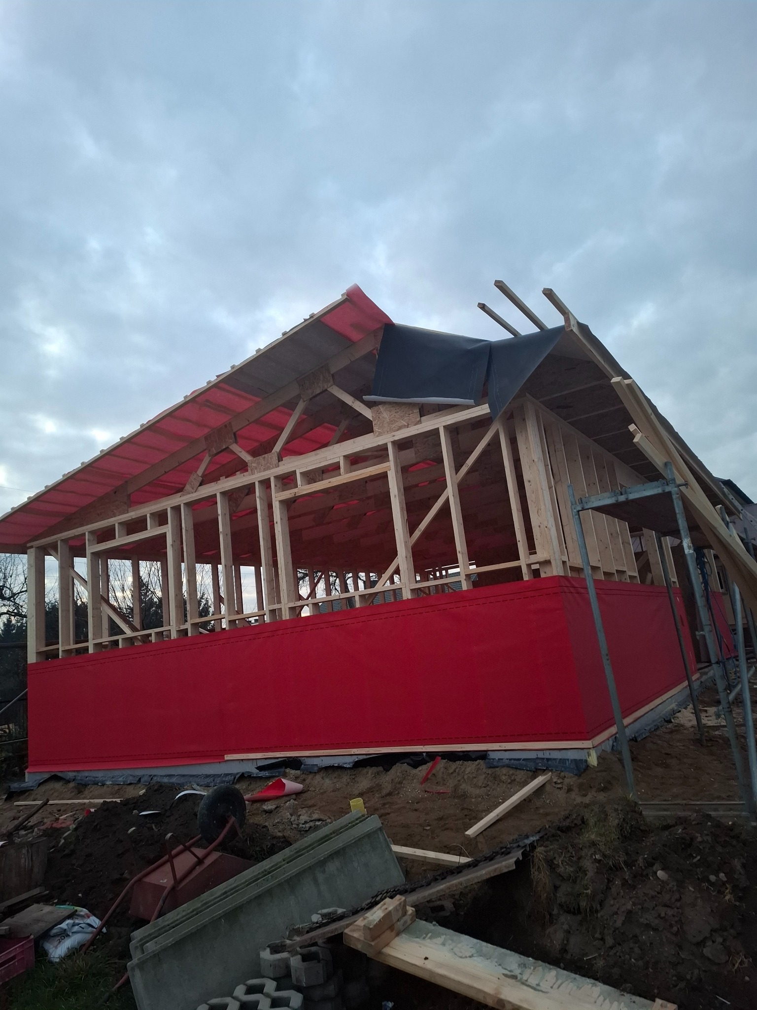 Konstrukcja drewniana domu w trakcie budowy, widoczna więźba dachowa z częściowo rozłożoną membraną, czerwone panele na ścianach szczytowych, pochmurne niebo w tle, widoczne narzędzia i materiały...