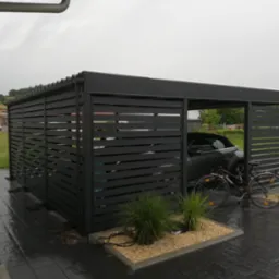 Nowoczesna, czarna pergola metalowa z poziomymi listwami, osłaniająca samochód i rower, z dekoracyjnymi trawami na żwirowym podłożu, na tle zieleni.