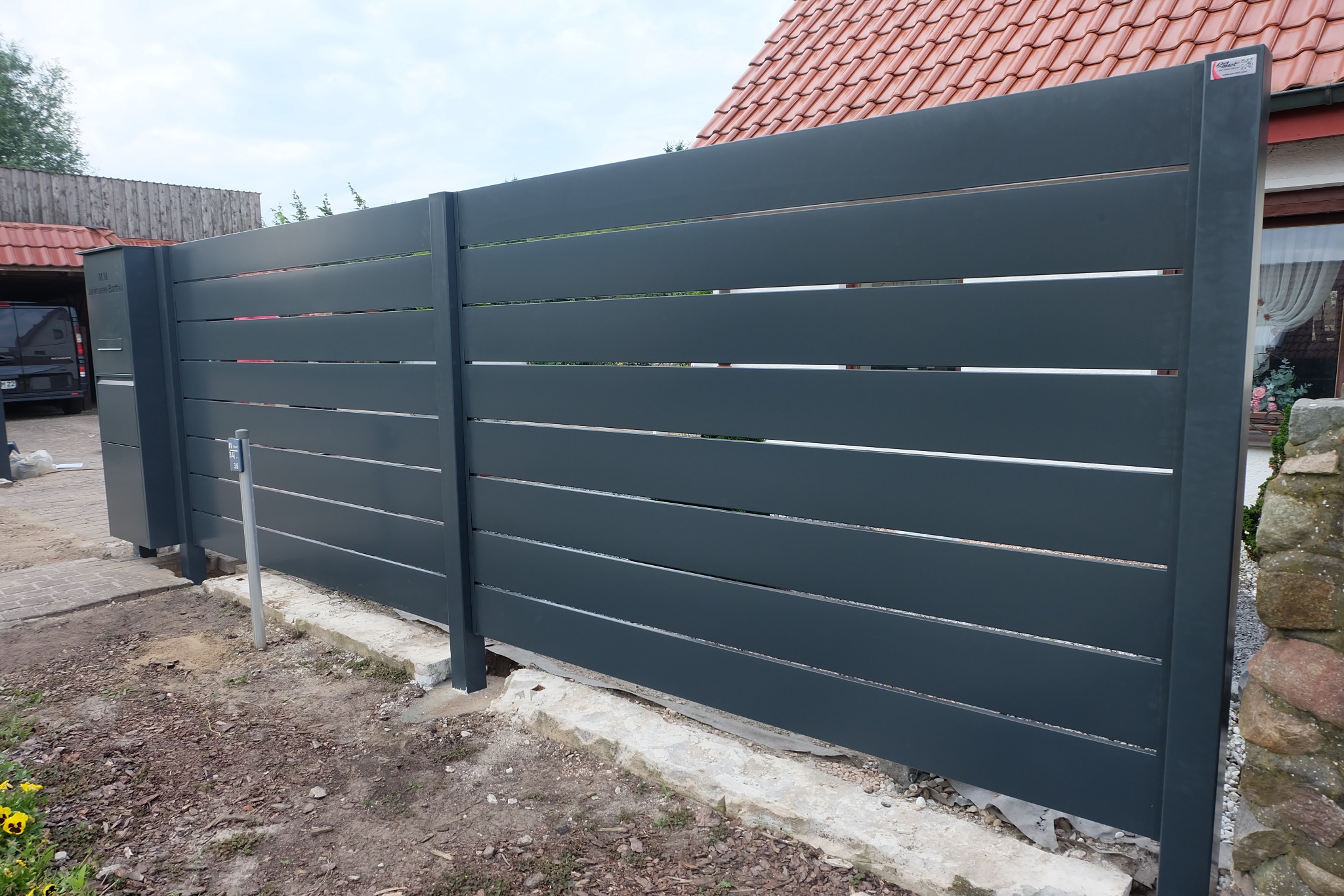 Nowoczesne, poziome, szare przęsła ogrodzeniowe zamontowane przy posesji z widocznym domem z czerwoną dachówką i fragmentem kamiennego murku.