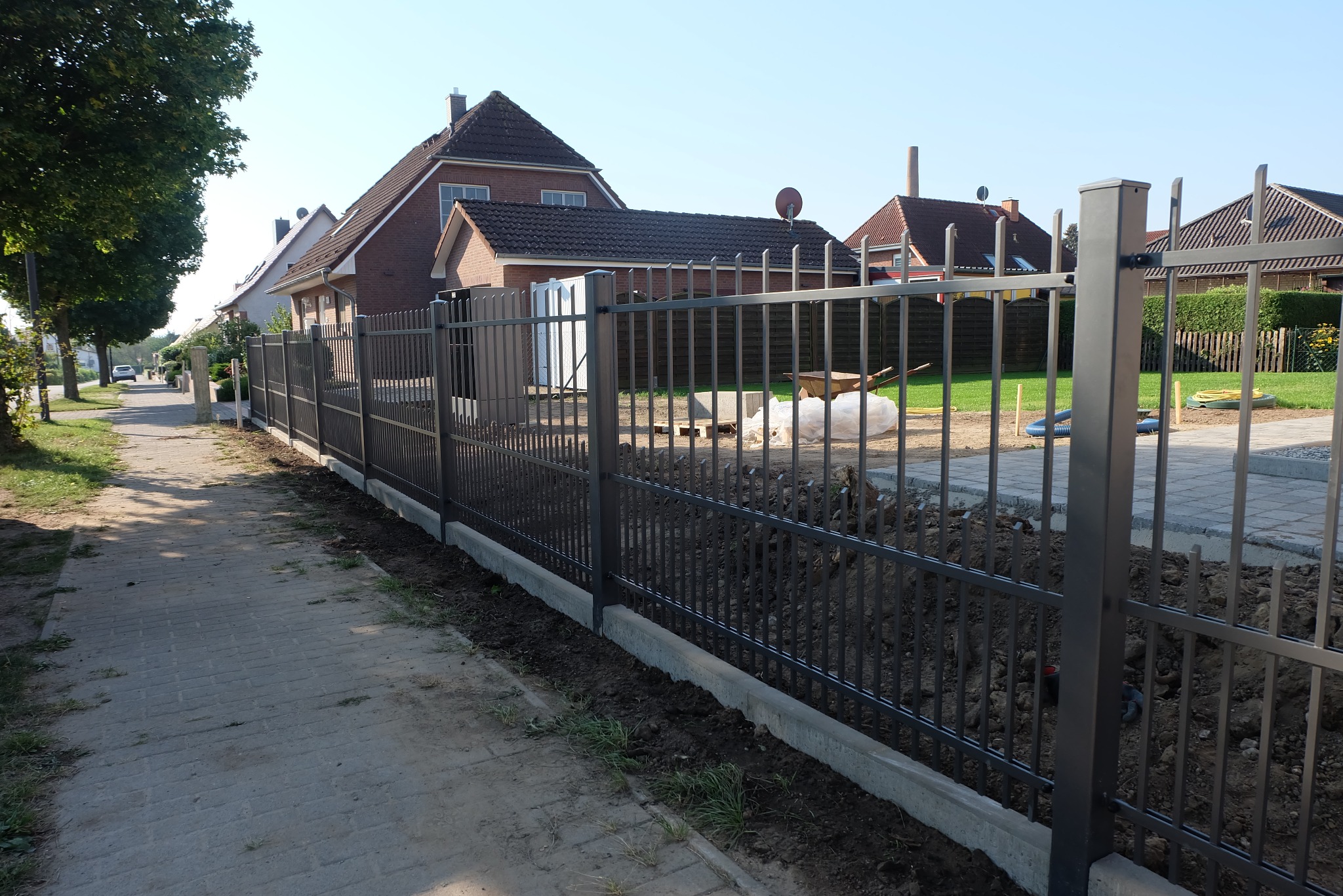 Nowoczesne, grafitowe ogrodzenie metalowe z pionowymi prętami, osadzone na betonowej podmurówce, oddzielające posesję od chodnika z kostki brukowej; w tle domy jednorodzinne.