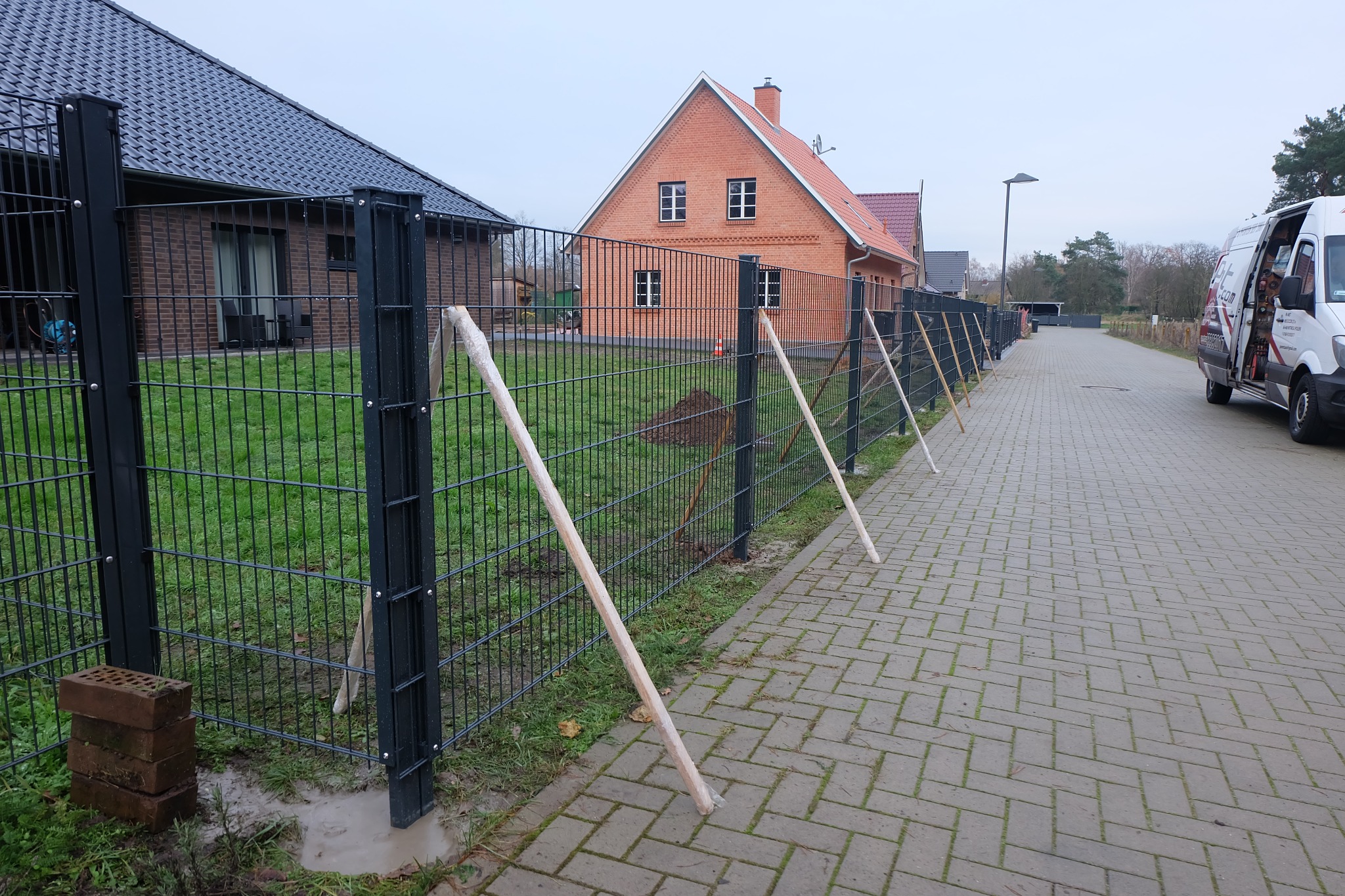 Montaż czarnego ogrodzenia panelowego z podporami tymczasowymi wzdłuż chodnika, w tle domy z cegły i samochód dostawczy z otwartymi drzwiami.