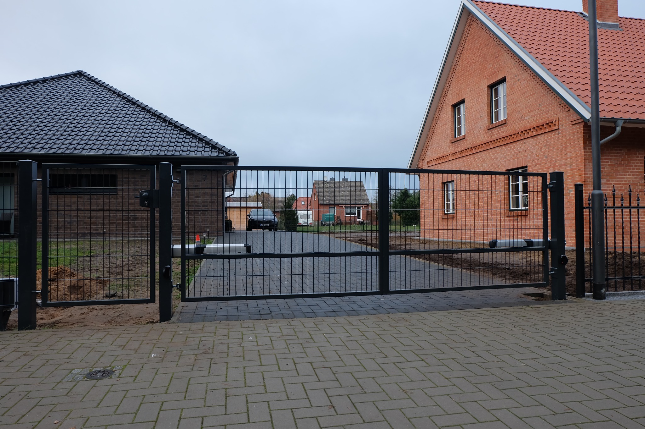 Nowoczesna, automatyczna brama wjazdowa panelowa w kolorze grafitowym, z widocznymi siłownikami, na tle dwóch domów z czerwonej cegły i kostki brukowej.