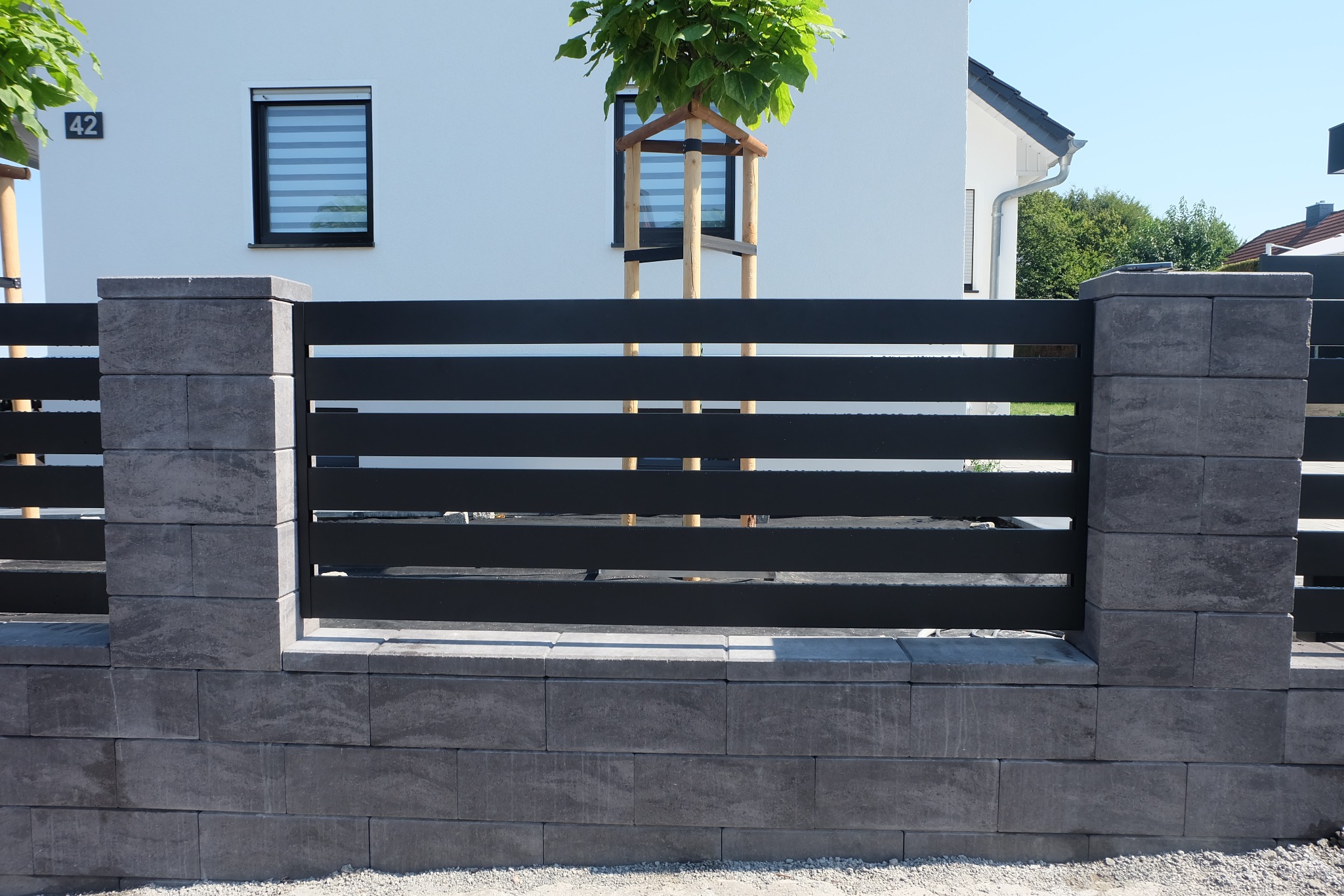 Nowoczesne ogrodzenie panelowe z poziomych, czarnych listew i szarych betonowych słupków, na tle białego domu z numerem 42 i młodym drzewem.