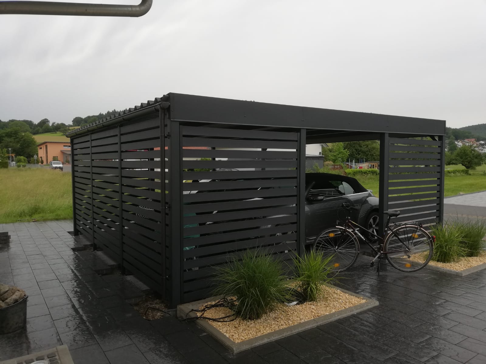 Nowoczesna, czarna pergola metalowa z poziomymi listwami, osłaniająca samochód i rower, z dekoracyjnymi trawami na żwirowym podłożu, na tle zieleni.