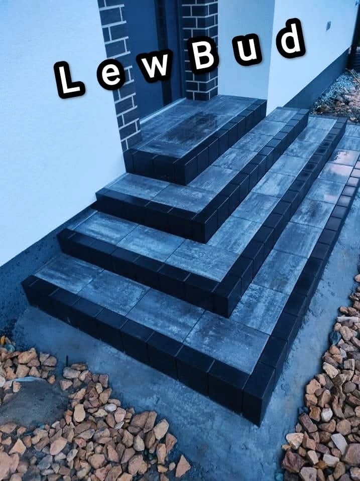 Schody wejściowe wyłożone szarą kostką brukową z czarnym obramowaniem, prowadzące do drzwi wejściowych; obok schodów widoczne kamienie ozdobne.