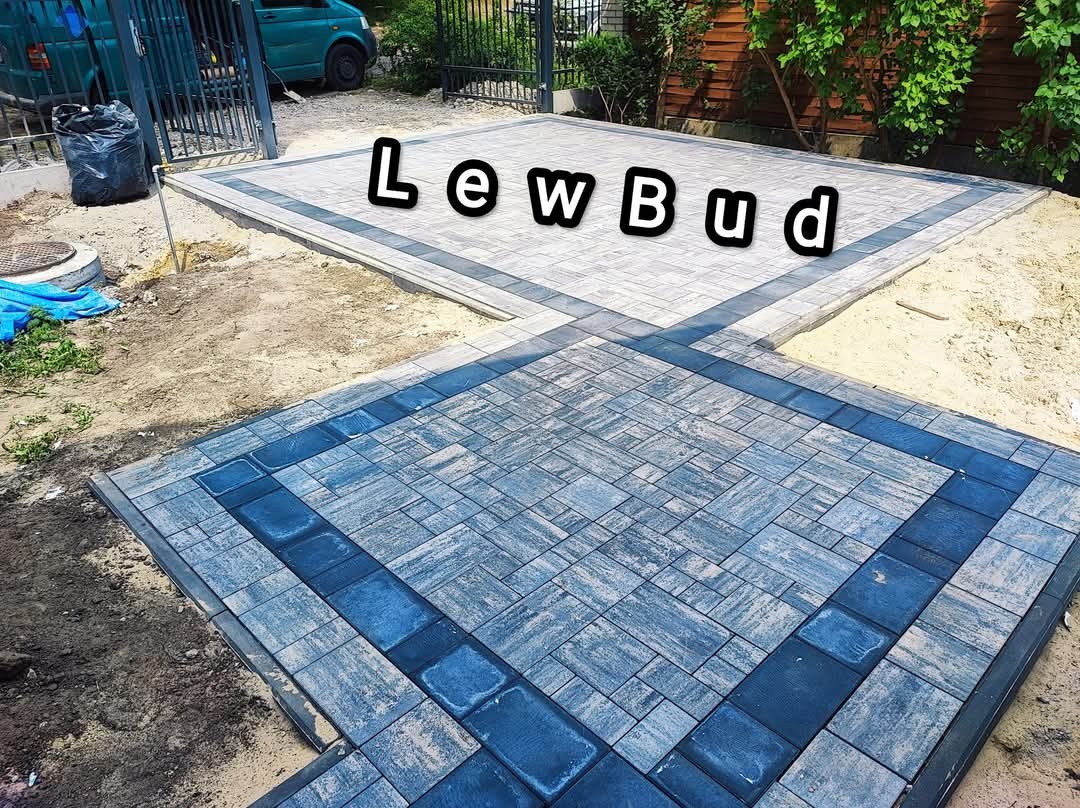 Ułożona kostka brukowa w dwóch odcieniach szarości z ciemnoniebieskimi obramowaniami, widoczna część posesji z piaskiem i trawą.