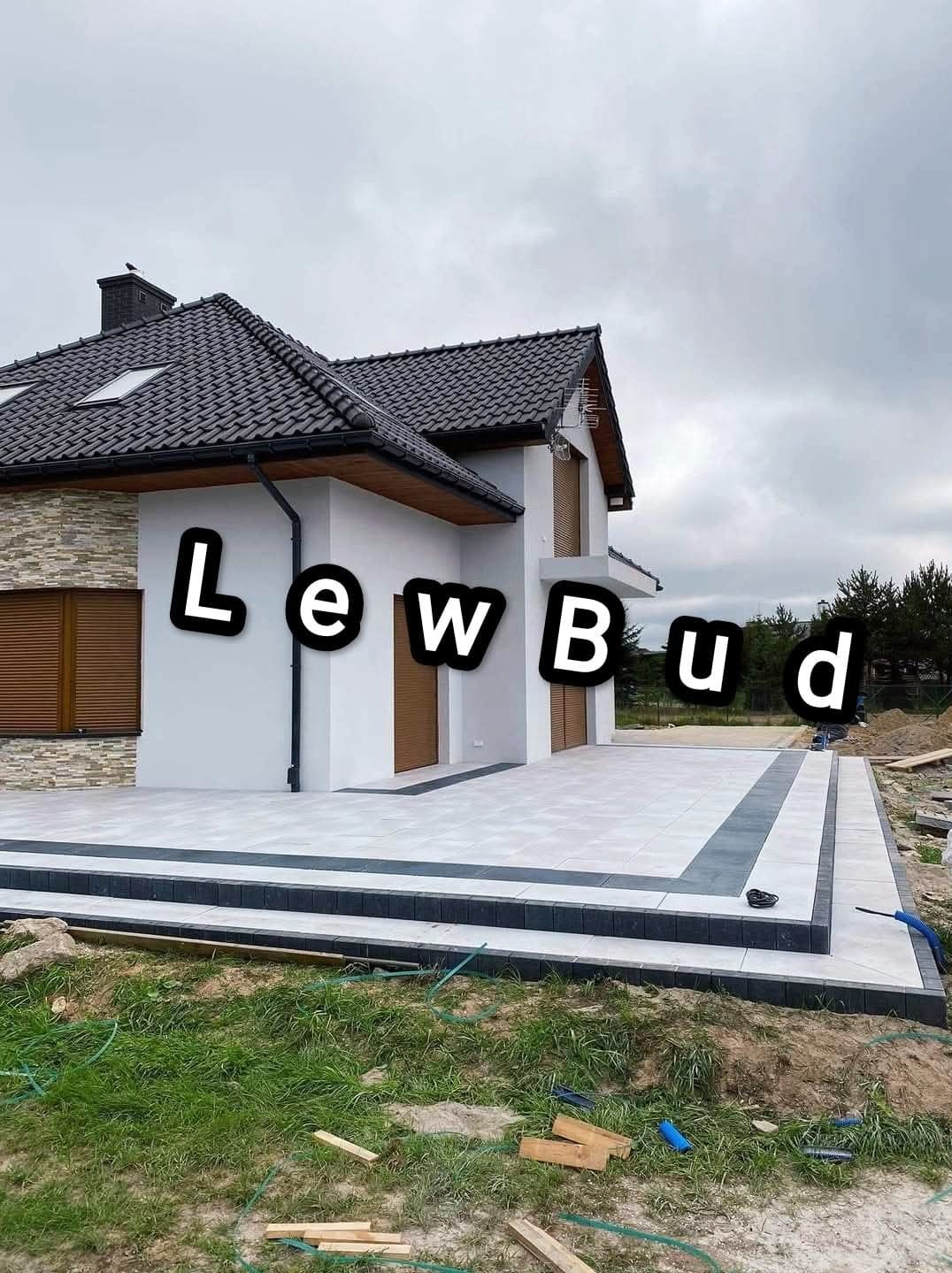 Nowoczesny dom z tarasem wyłożonym jasnymi płytami, obrzeżonymi ciemniejszą kostką. Na pierwszym planie trawnik z drewnianymi elementami budowlanymi.