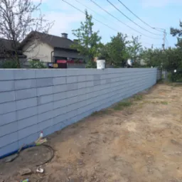 Nowo postawiony, szary mur z betonowych bloczk&oacute;w, widok z zewnątrz posesji, z widocznymi narzędziami i materiałami budowlanymi przy podstawie. W tle domy i drzewa.