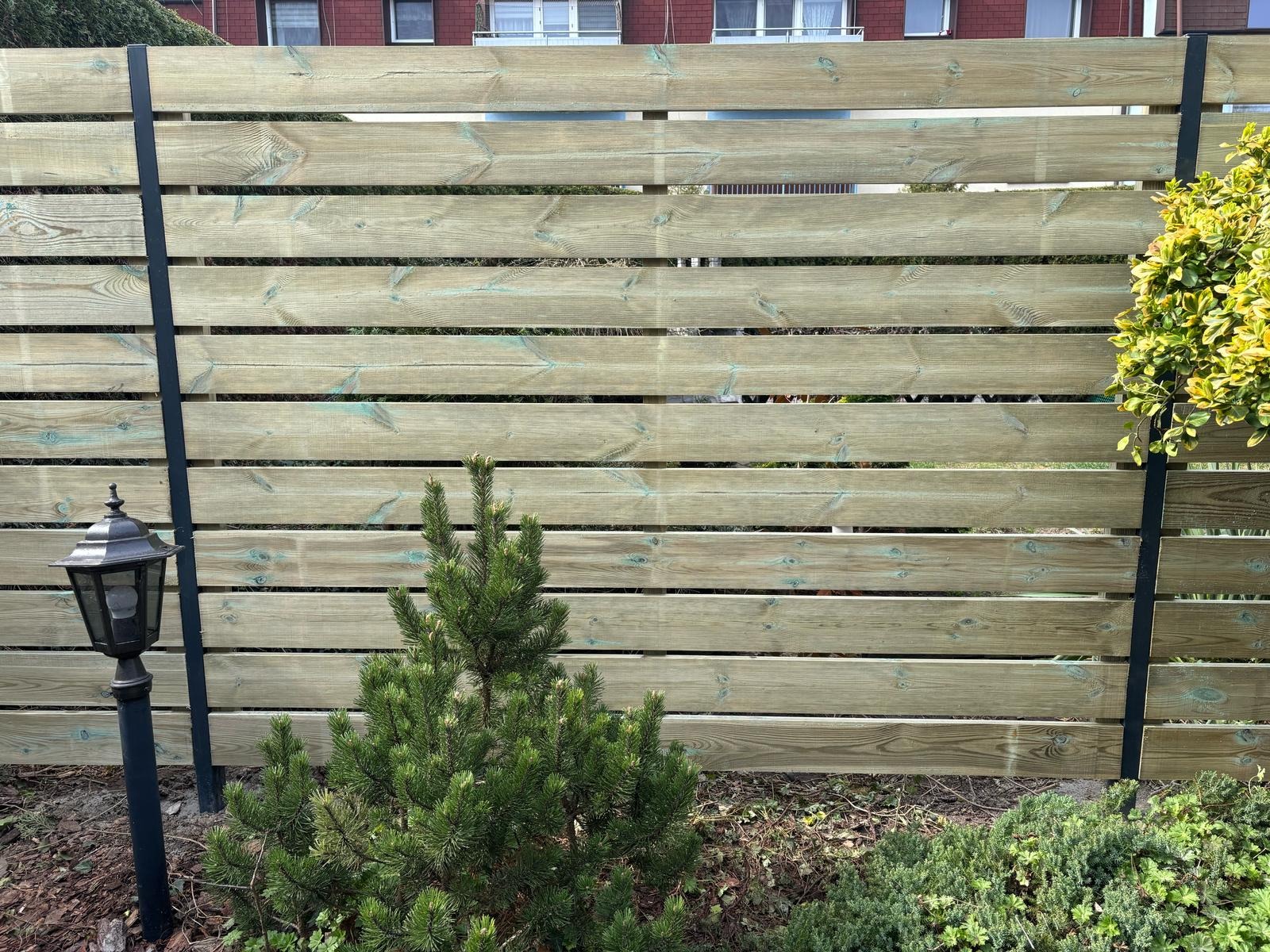 Nowoczesne ogrodzenie poziome z drewnianych desek, wsparte na metalowych słupkach, z lampą ogrodową i iglakiem na pierwszym planie.