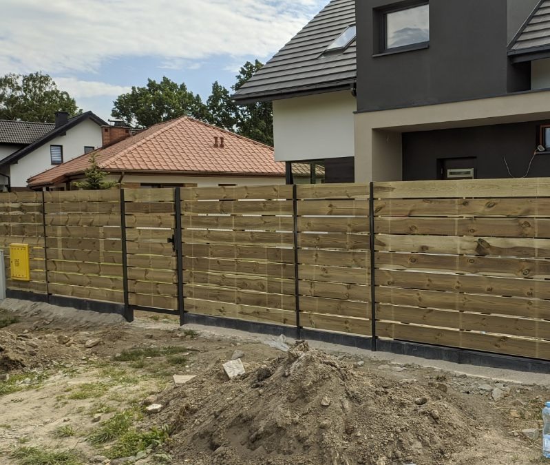 Nowoczesne ogrodzenie poziome z jasnego drewna, z czarnymi słupkami i furtką, na tle dwóch domów o różnej architekturze, widoczny stos ziemi przed ogrodzeniem.