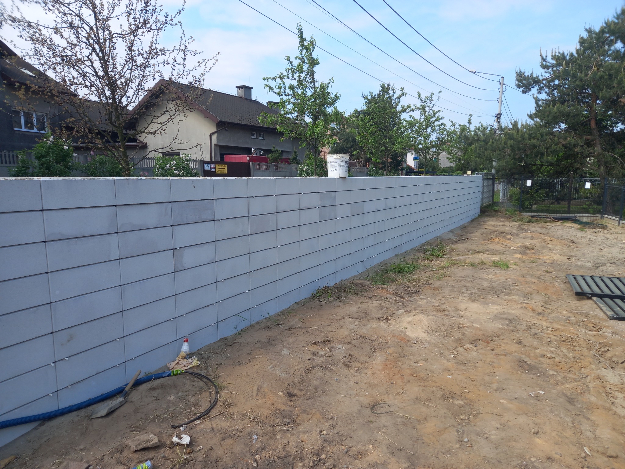 Nowo postawiony, szary mur z betonowych bloczków, widok z zewnątrz posesji, z widocznymi narzędziami i materiałami budowlanymi przy podstawie. W tle domy i drzewa.