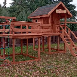 Walczak - Kompleksowy plac zabaw z drewna: domek na podeście ze schodami, huśtawka i ścianka wspinaczkowa z kolorową siatką, otoczony zielenią. Tabliczka 'Sprzedam' na domku.
