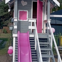Walczak - Uroczy, szary domek zabaw dla dzieci z różową zjeżdżalnią i akcentami, ozdobiony sercem i lampkami, otoczony zielenią. Otwarta, różowa furtka zaprasza do środka.