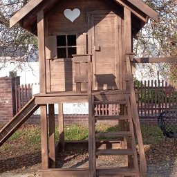 Walczak - Drewniany domek zabaw dla dzieci z schodkami i zjeżdżalnią, z serduszkiem wyciętym w ściance, na tle drzew i ogrodzenia. Całość w ciepłym, brązowym odcieniu.