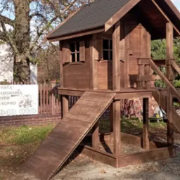 Drewniany domek dla dzieci z zadaszeniem i zjeżdżalnią, w odcieniach brązu, umieszczony na trawniku obok drzewa z brązowymi liśćmi i białej tablicy z napisem.