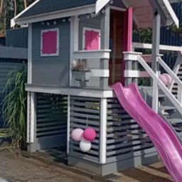 Drewniany domek dla dzieci w szaro-białej kolorystyce z różowymi akcentami, wyposażony w zjeżdżalnię i ozdobiony lampionami oraz balonami. Stoi na piaszczystym podłożu.