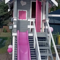 Szara altanka dla dzieci z różową zjeżdżalnią i otwieranymi drzwiami, ozdobiona sercem i lampkami, umieszczona w ogrodzie.