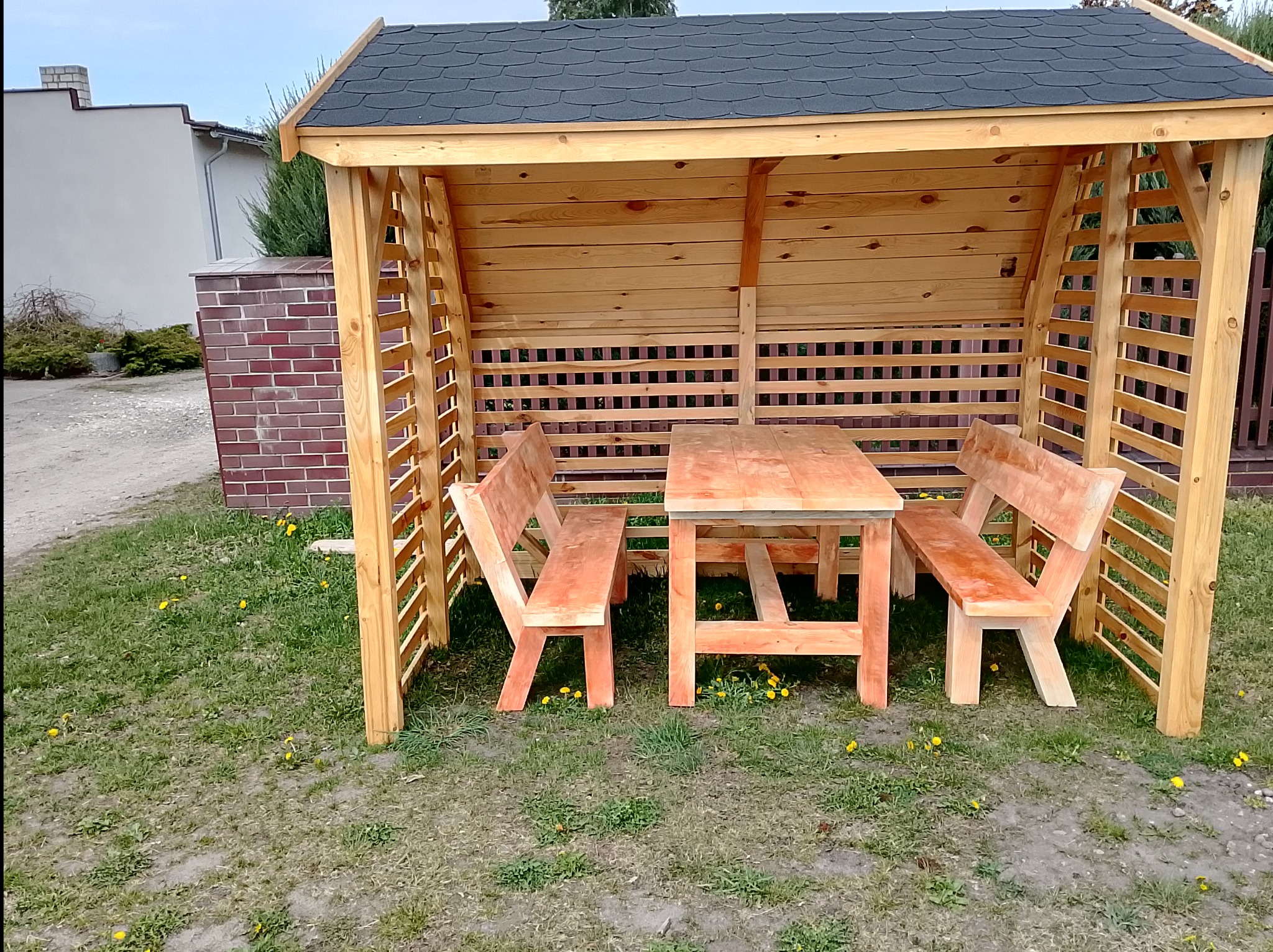 Drewniana altana ogrodowa z zadaszeniem, stół i dwie ławki w środku, na trawie. Konstrukcja z jasnego drewna, ciemny dach. Widok z przodu, w tle budynek.