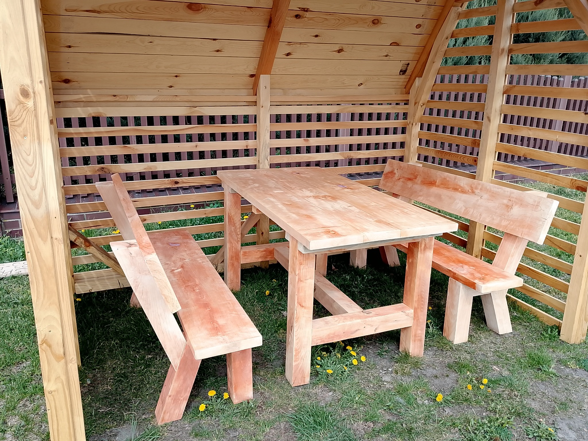 Drewniana altana z ażurowymi ścianami i meblami ogrodowymi: stół i dwie ławki z oparciami, całość w naturalnym odcieniu drewna, na tle zielonego trawnika.