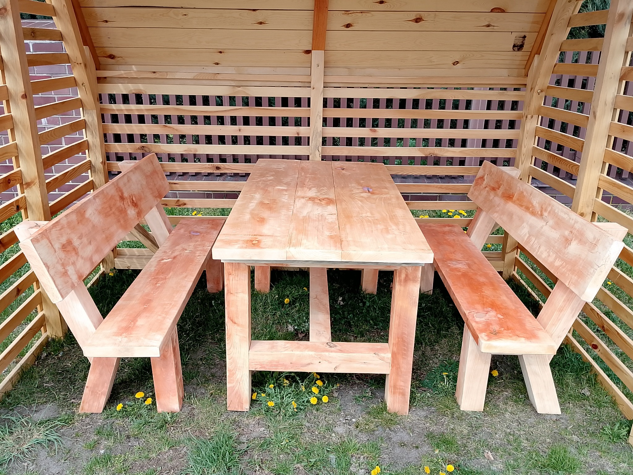 Drewniana altana z prostym stołem i ławkami, wykonana z jasnego drewna, otoczona zielonym trawnikiem z żółtymi kwiatami. Widok z przodu, oświetlenie dzienne.