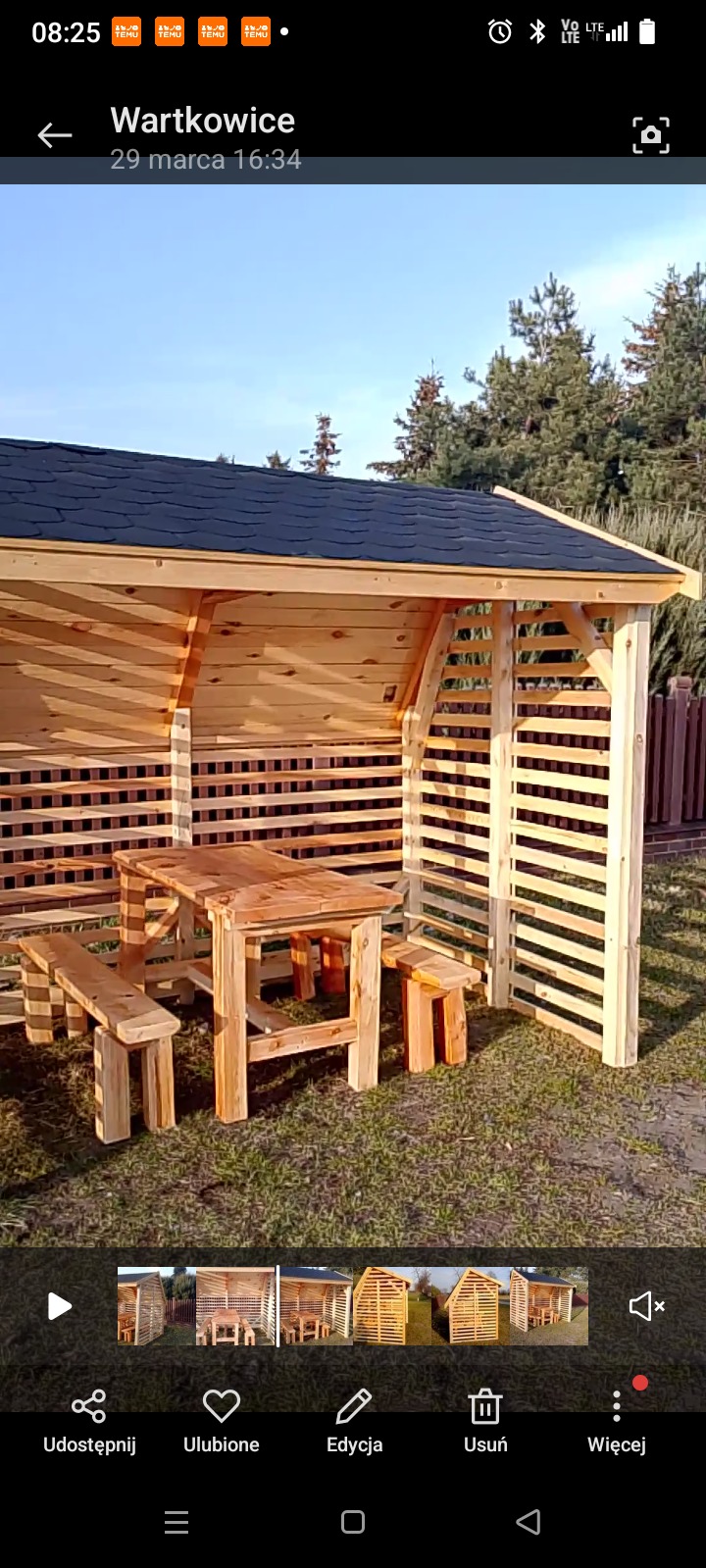 Drewniana altana z czarnym dachem, wyposażona w stół i ławki z surowego drewna, ustawiona na trawniku. Konstrukcja z ażurowymi ścianami, widok na zielone drzewa w tle.