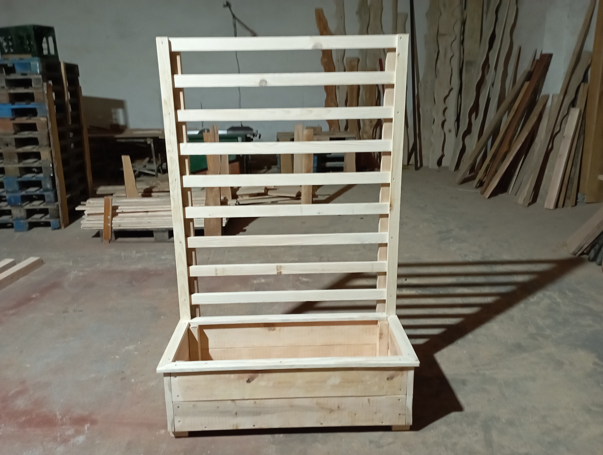 Surowa, drewniana konstrukcja z donicą i kratką, widoczna w warsztacie stolarskim. Jasne drewno, prosta forma, bez wykończenia. Tło z palet i desek.