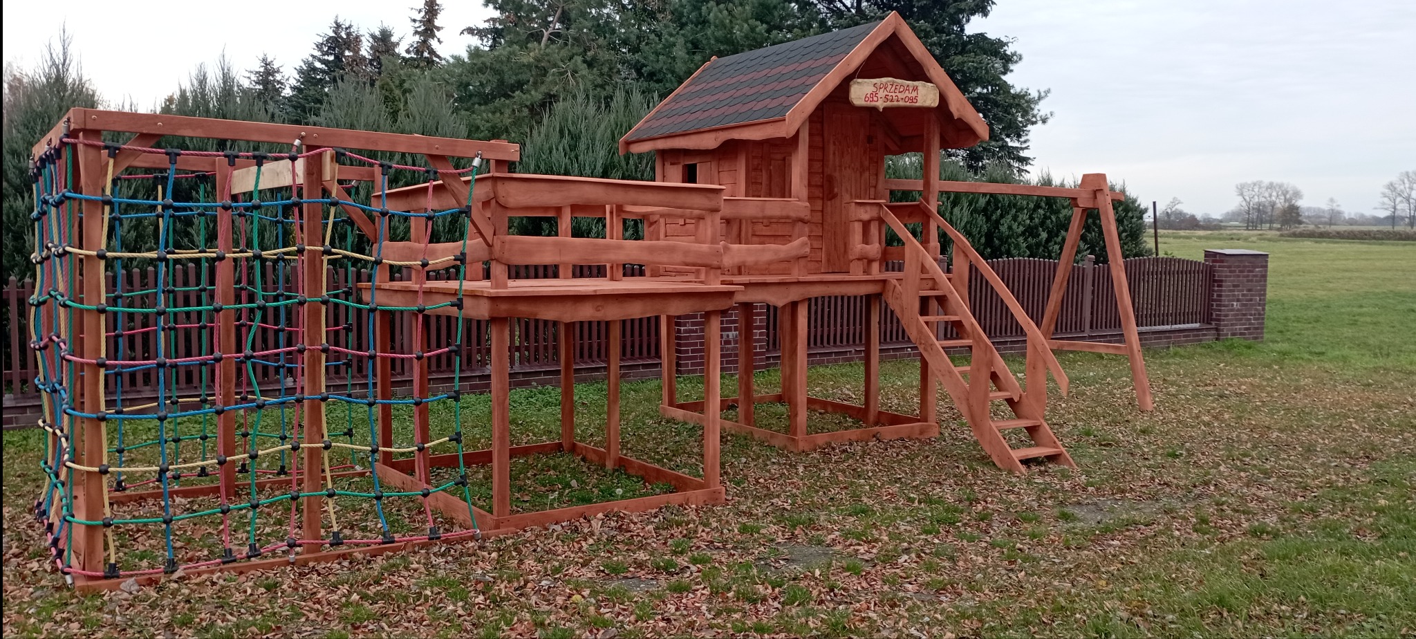 Kompleksowy plac zabaw z drewna: domek na podeście ze schodami, huśtawka i ścianka wspinaczkowa z kolorową siatką, otoczony zielenią. Tabliczka 'Sprzedam' na domku.