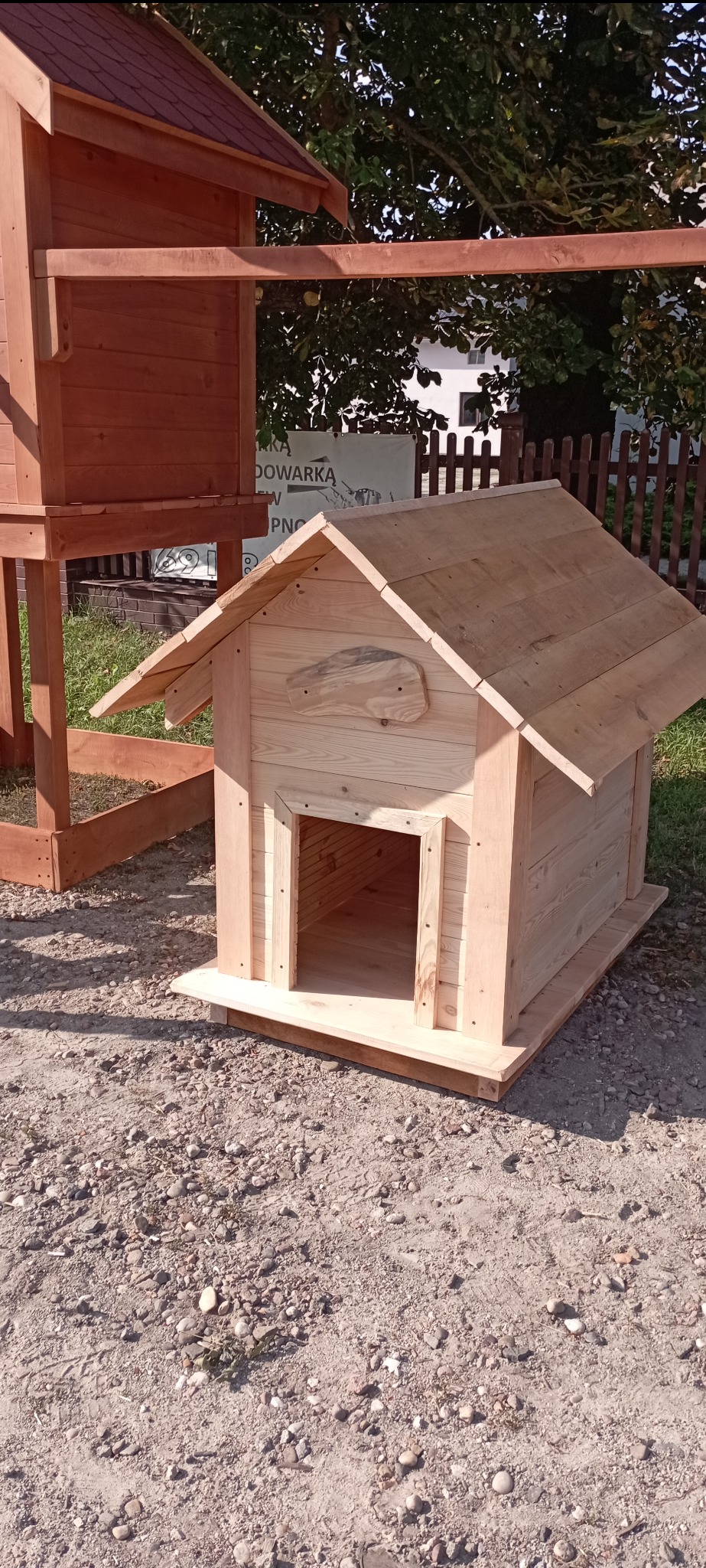 Drewniana buda dla psa z surowego drewna, z charakterystyczną ozdobą w kształcie mapy na ścianie, na tle częściowo widocznej drewnianej konstrukcji i zieleni.