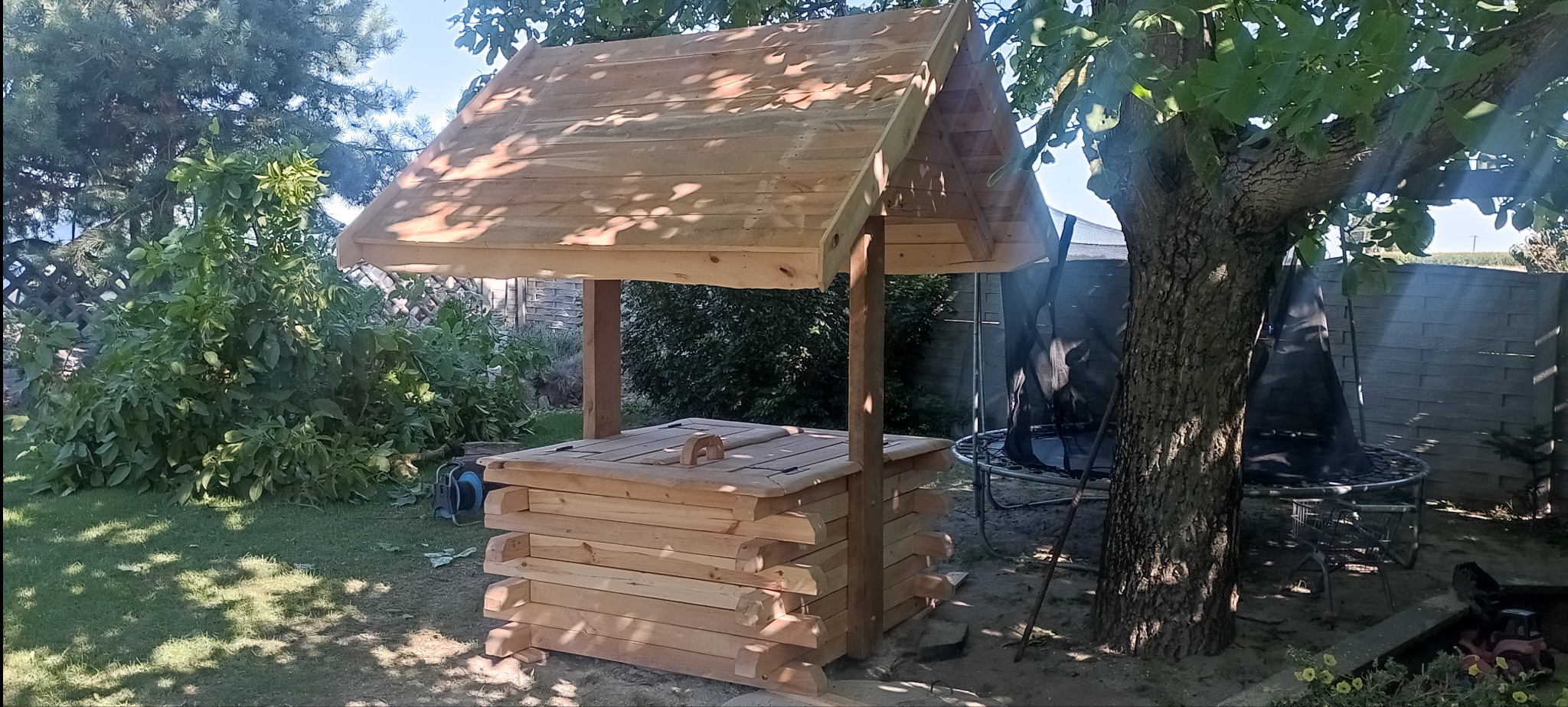 Drewniana studnia ogrodowa z dachem, wykonana z jasnego drewna, umieszczona na trawniku w ogrodzie, w tle drzewa i ogrodzenie.