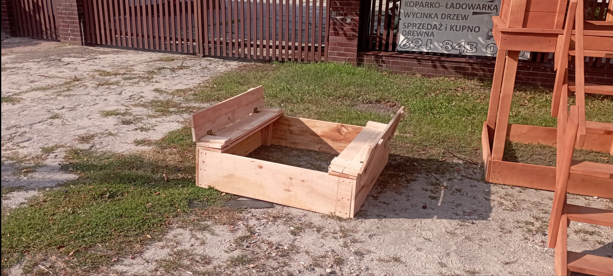 Surowa, drewniana konstrukcja, prawdopodobnie skrzynia z siedziskiem, na trawiastym podłożu. W tle płot i budynek z reklamą. Widoczny fragment drabiny.