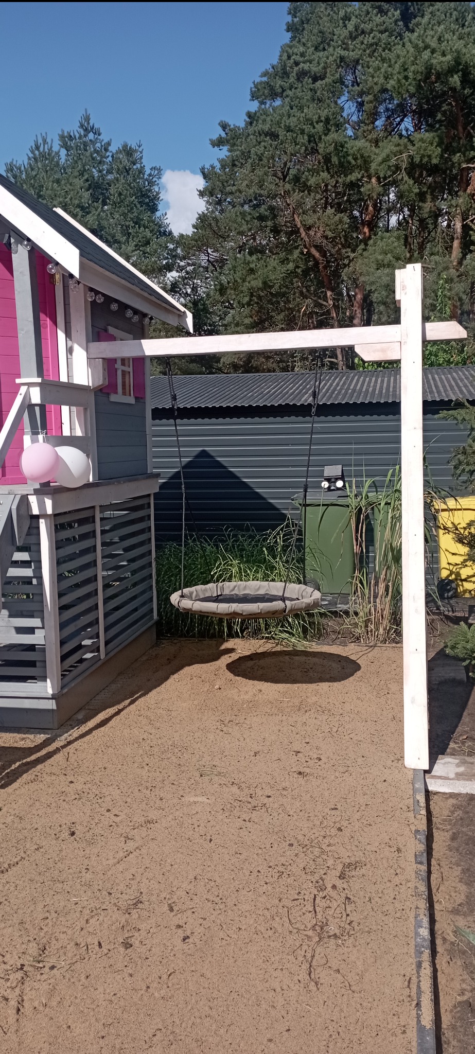 Kolorowa altana z huśtawką typu bocianie gniazdo na piaszczystym podłożu. Elementy drewniane, jasne światło słoneczne.