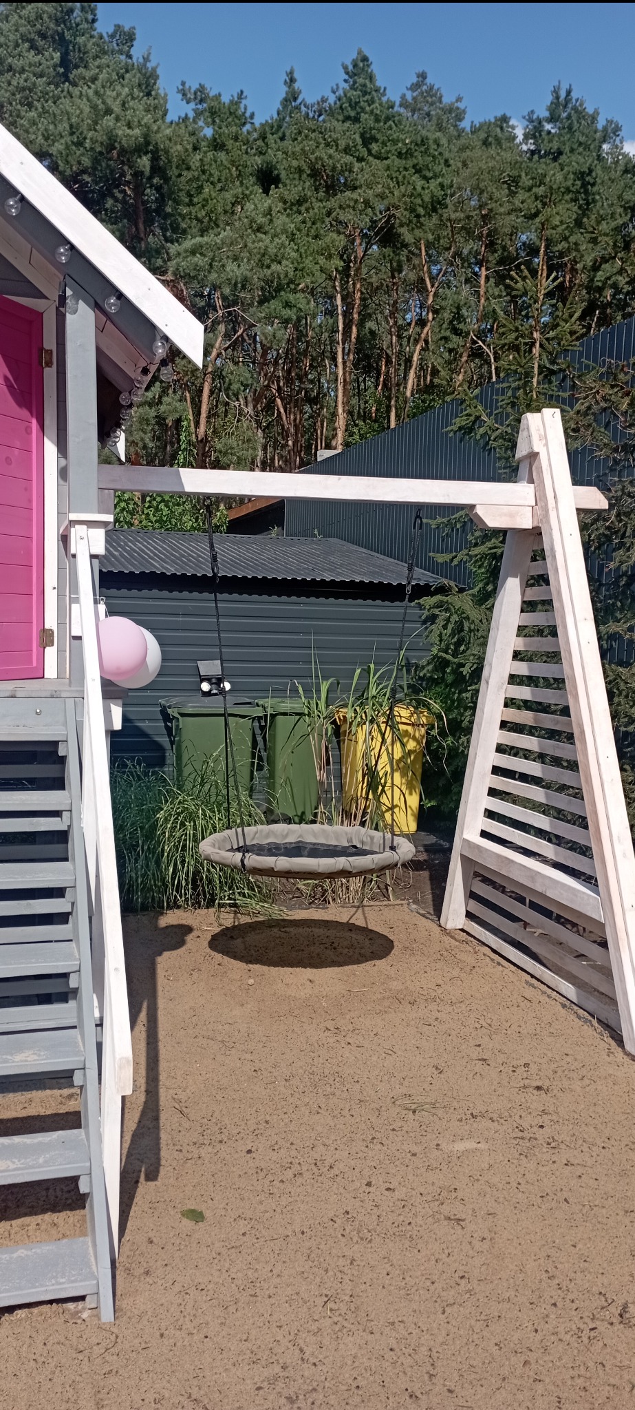 Drewniana huśtawka typu gniazdo zawieszona na konstrukcji z jasnego drewna. W tle widoczny domek z różowymi drzwiami, zieleń i szare pojemniki na śmieci.