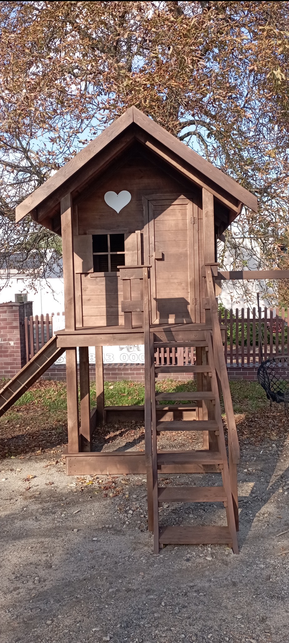 Drewniany domek zabaw dla dzieci z schodkami i zjeżdżalnią, z serduszkiem wyciętym w ściance, na tle drzew i ogrodzenia. Całość w ciepłym, brązowym odcieniu.